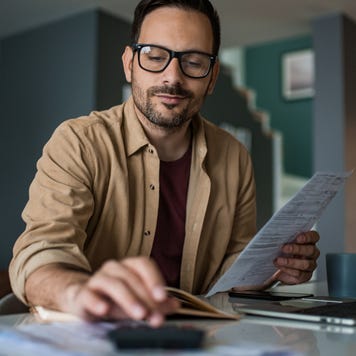 Young man working on finances at home. He has a laptop open and is using a calculator.