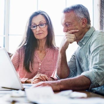 Senior couple sitting at a dining table, using a laptop computer, paperwork and bills on the table
