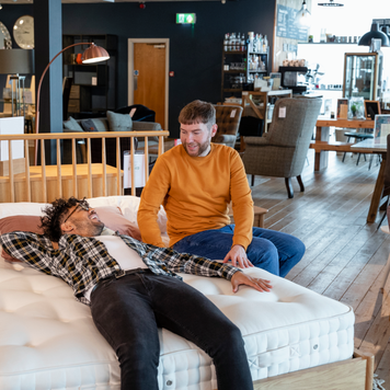 A mid adult couple viewing a home furniture showroom together, one man is lying on a bed while his fiancé sits near him on the bed. They are testing out the bed and considering buying it.